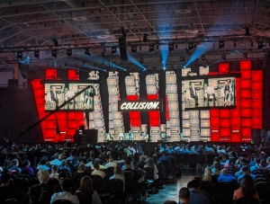 Image of centre stage at tech conference with stages and screens and crowd in attendance. 